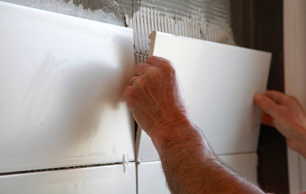 Pose de carrelage mural : installation de carreaux blancs Mains d'un ouvrier posant une nouvelle tuile murale blanche et brillante sur un mortier frais avec des croisillons d'espacement.