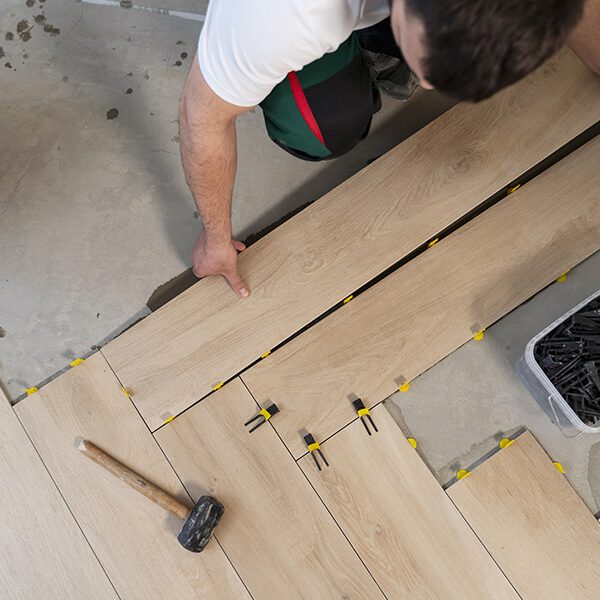 Pose carrelage imitation bois : artisan à l'ouvrage Ouvrier posant des carreaux imitation parquet bois clair sur sol en béton avec croisillons jaunes et noirs, utilisant un maillet.