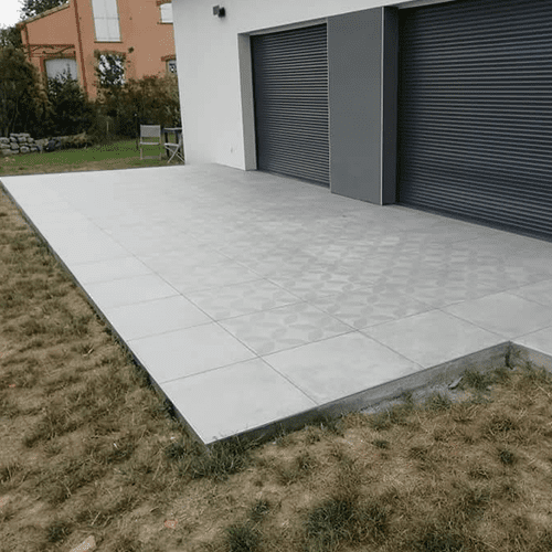 Terrasse Carrelage Extérieur Moderne et Jardin Grande terrasse en dalles de grès cérame gris clair à motifs, juxtaposée à une façade moderne blanche et portes volets roulants gris foncé.