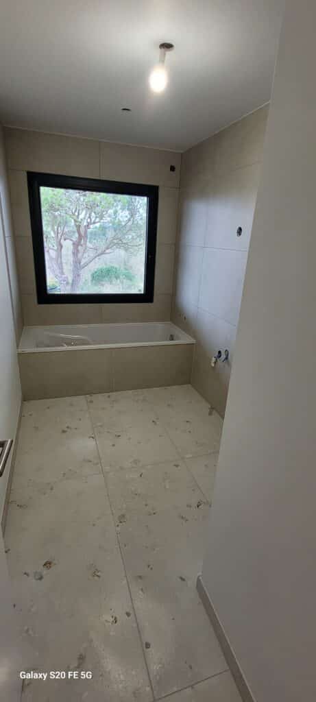 Salle de bain avec baignoire, grande fenêtre noire sur jardin et carrelage beige moderne.